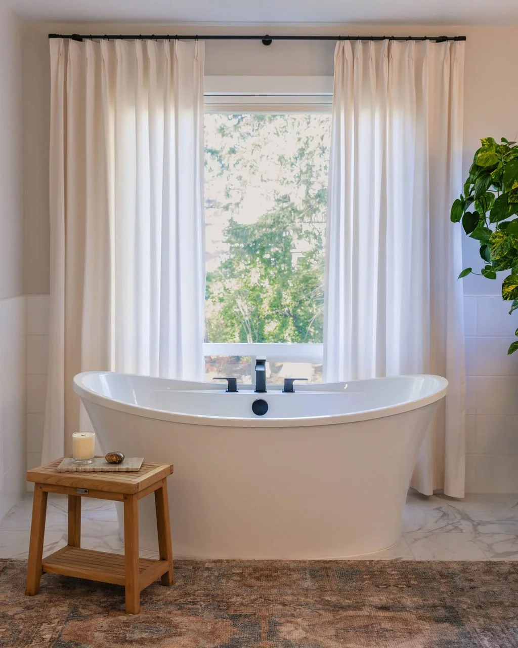 Primary bathroom with a freestanding soaking tub in front of a window with white drapery, a small wood stool, and light marble tile for a bright, softened feel.