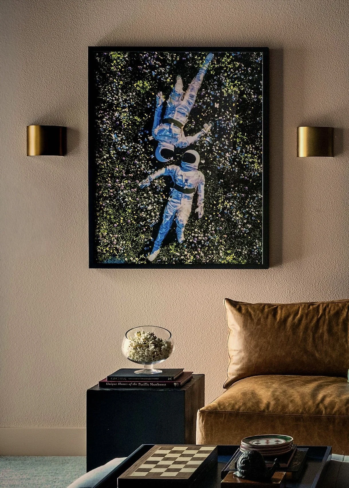 Framed astronaut artwork between two brass wall sconces above a small side table with decor, shown against a textured neutral wall.