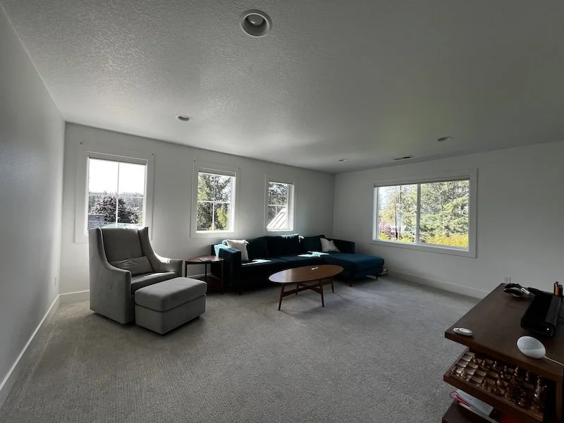 Spacious living room with large windows, light carpet, a dark sectional sofa, accent chairs, and a round coffee table, filled with natural light and open sightlines.