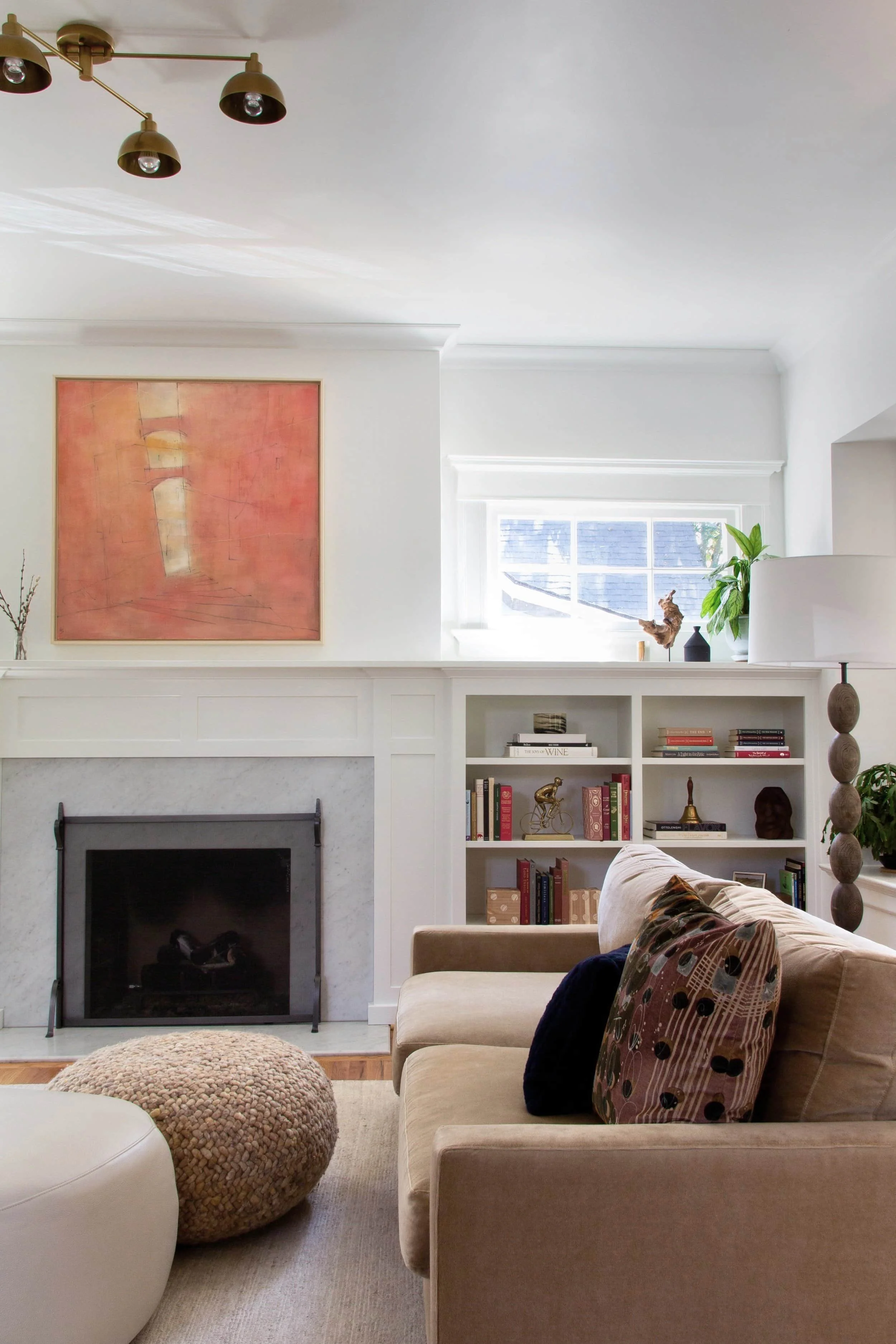 Living room with a beige couch and colorful throw pillows, a round pouf, a white built-in bookshelf, a fireplace with a marble surround, a window with plants and decorative items, and framed abstract art on the wall.