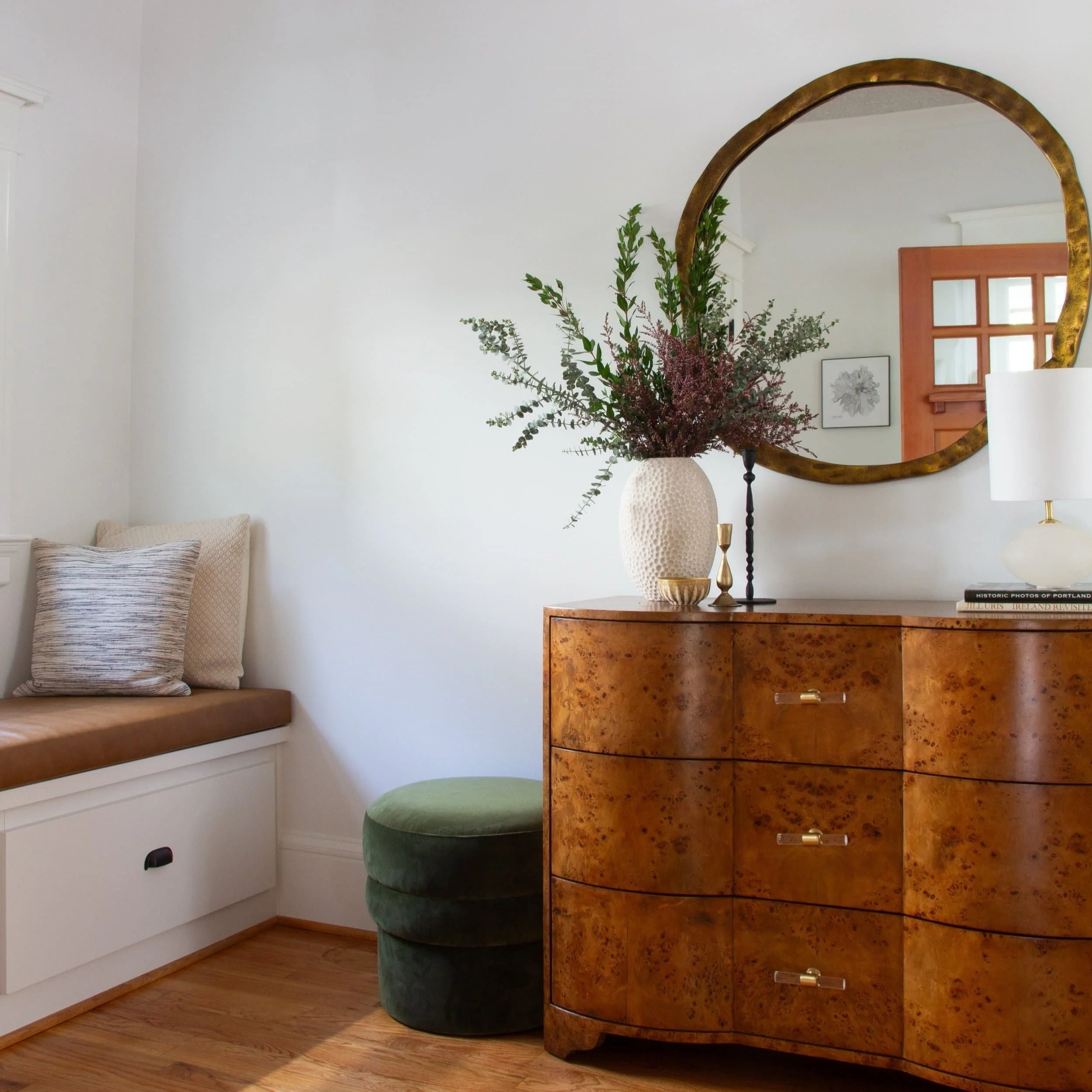 A foyer corner with a wooden dresser, a large mirror, a white vase with greenery, a white table lamp, and a window seat with pillows.