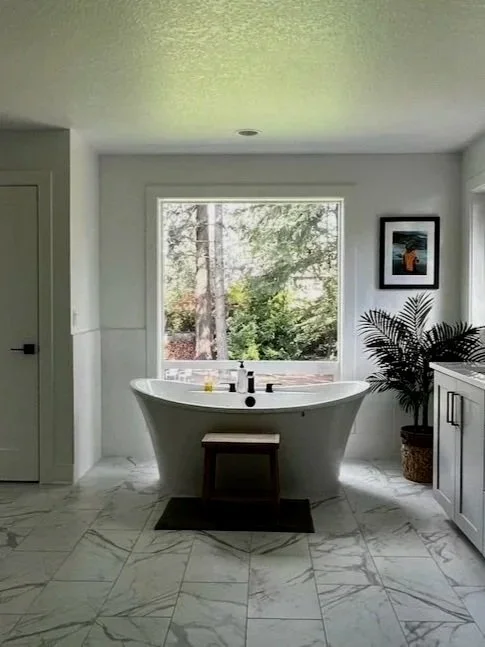 Bright bathroom featuring a freestanding soaking tub beneath a large window, marble tile floors, soft neutral walls, and simple modern finishes with natural light.