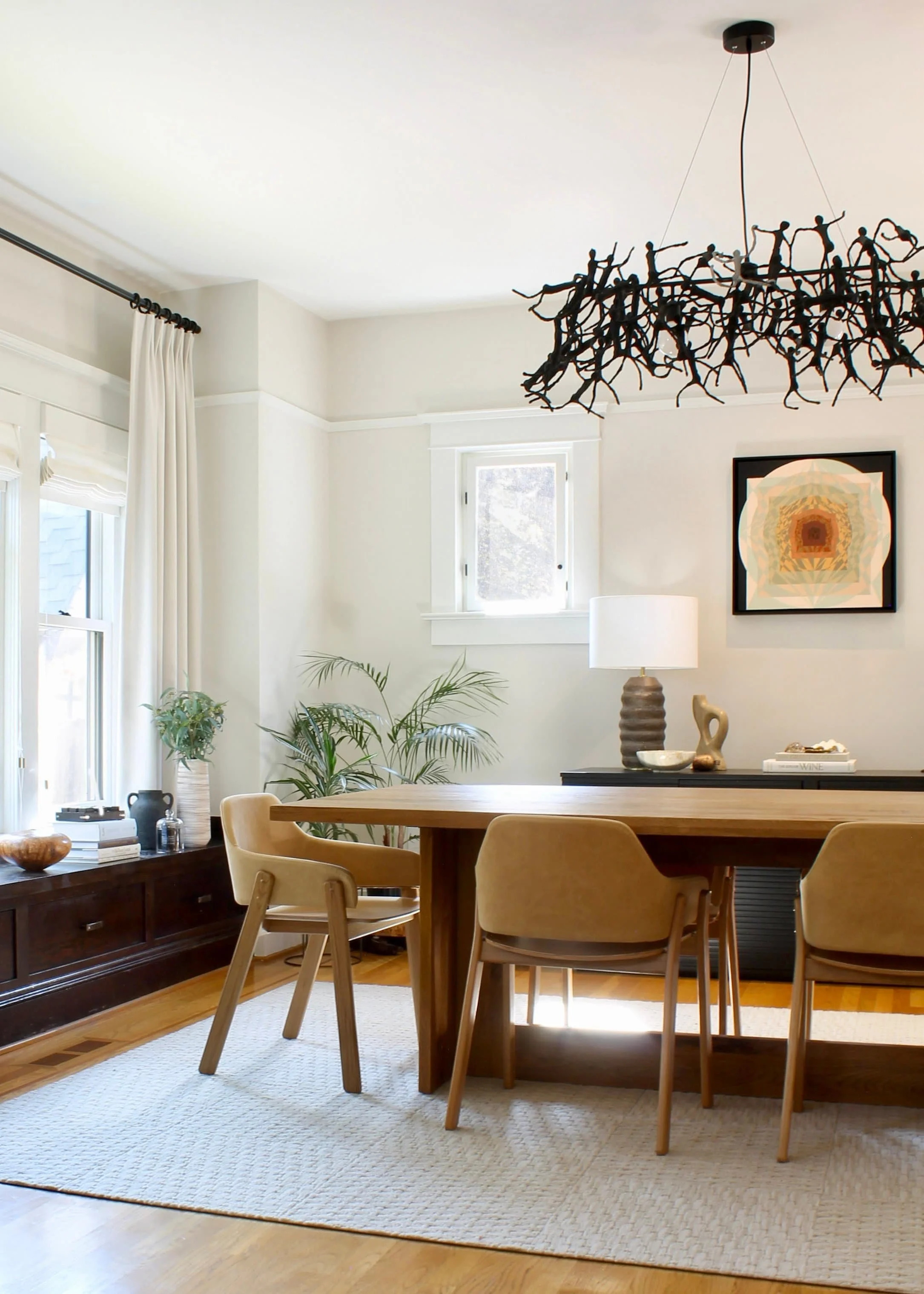 A modern dining room with a wooden table, beige chairs, and a large abstract chandelier. The room features light walls, a light rug, houseplants, and a piece of abstract art on the wall. A lamp and decorative items are on a side console.