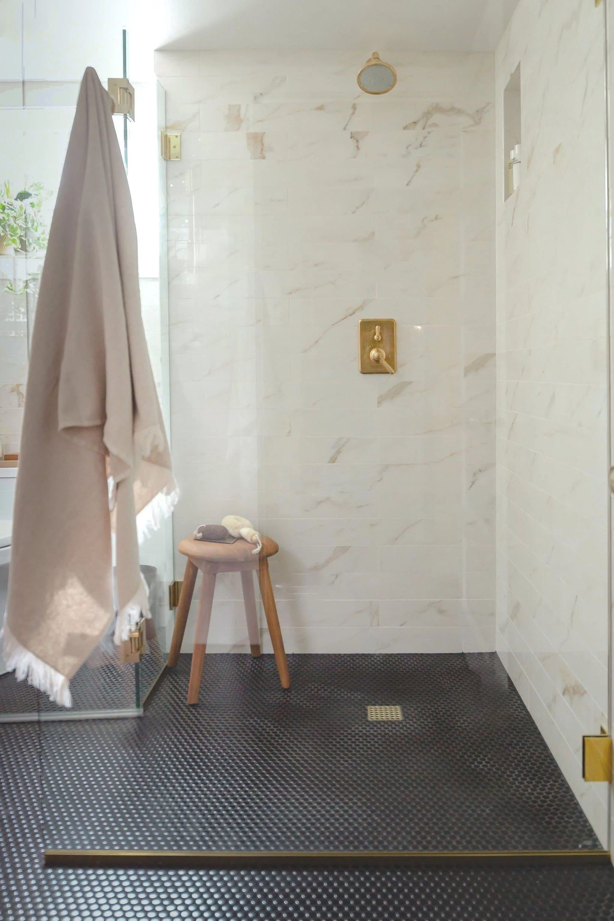 A modern bathroom shower area with light-colored marble tiles, a gold shower fixture, a small stool with rolled towels, and a glass door with a beige towel hanging on a hook.