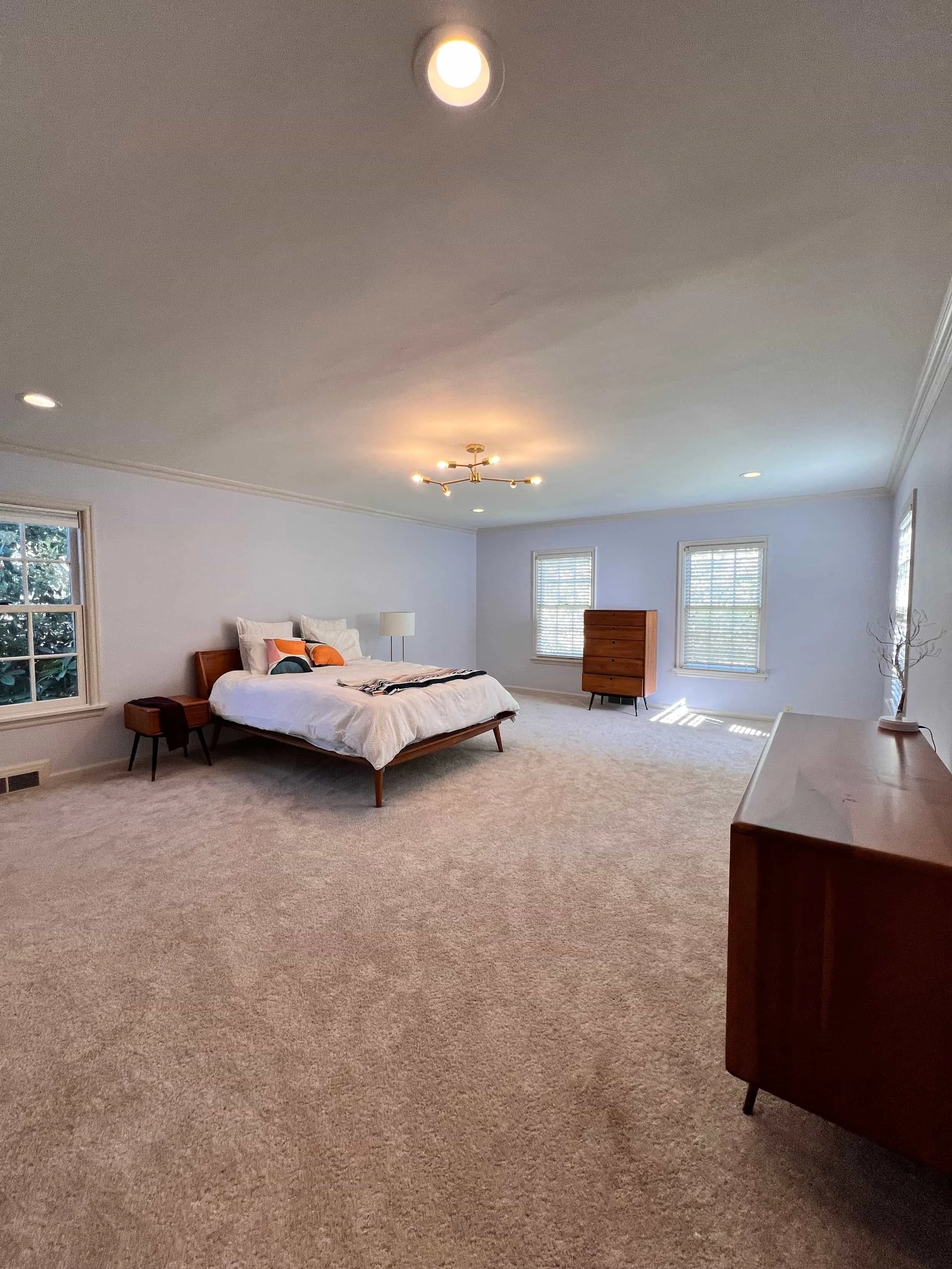A spacious bedroom with a bed, side tables, a dresser, and windows letting in natural light. The room has white walls, beige carpet, and simple modern furniture with a minimalistic design.