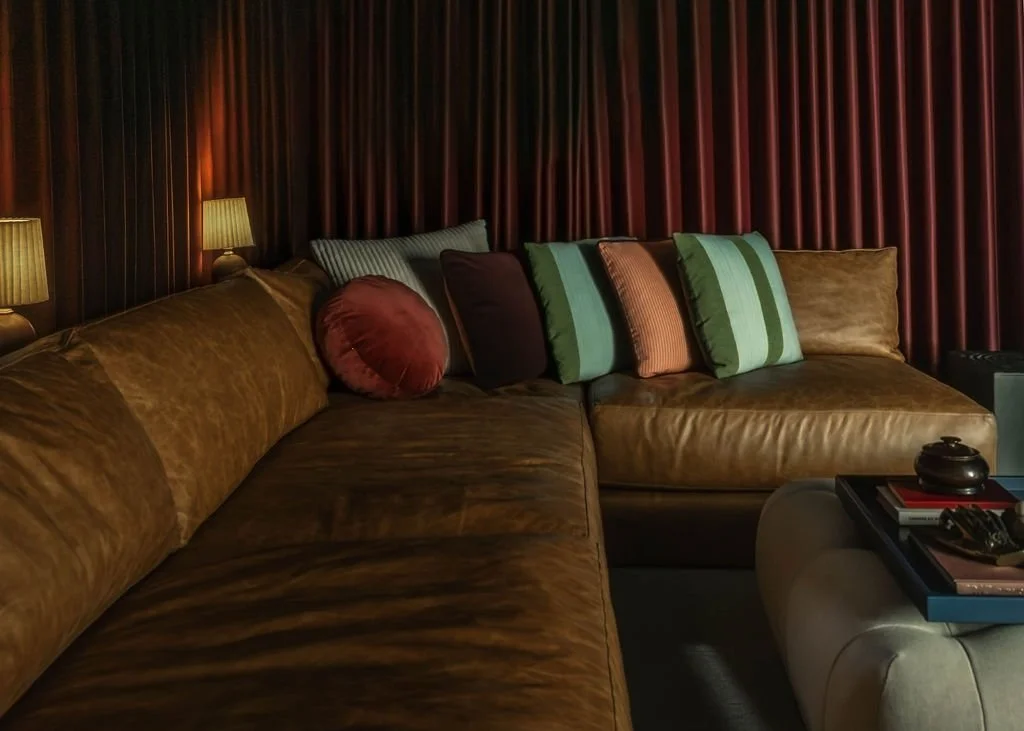 Close view of the media room sectional with layered pillows, warm table lamps, and wall to wall theater drapery creating a cozy, moody backdrop.