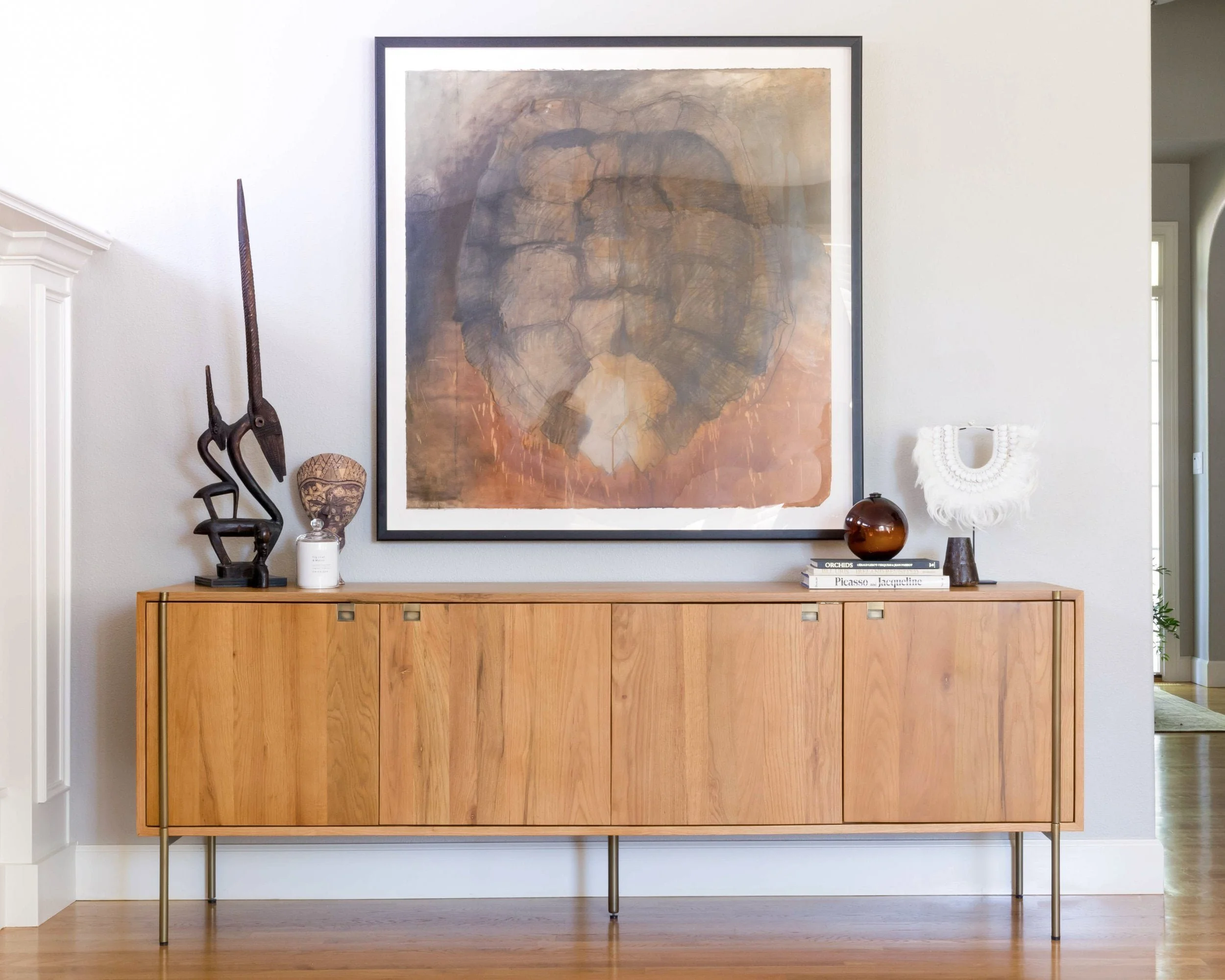 A modern interior setting featuring a wooden sideboard with decorative items like a sculptural piece, white jar, book stack, decorative bowl, and wall art. A large abstract painting is hung above the sideboard.