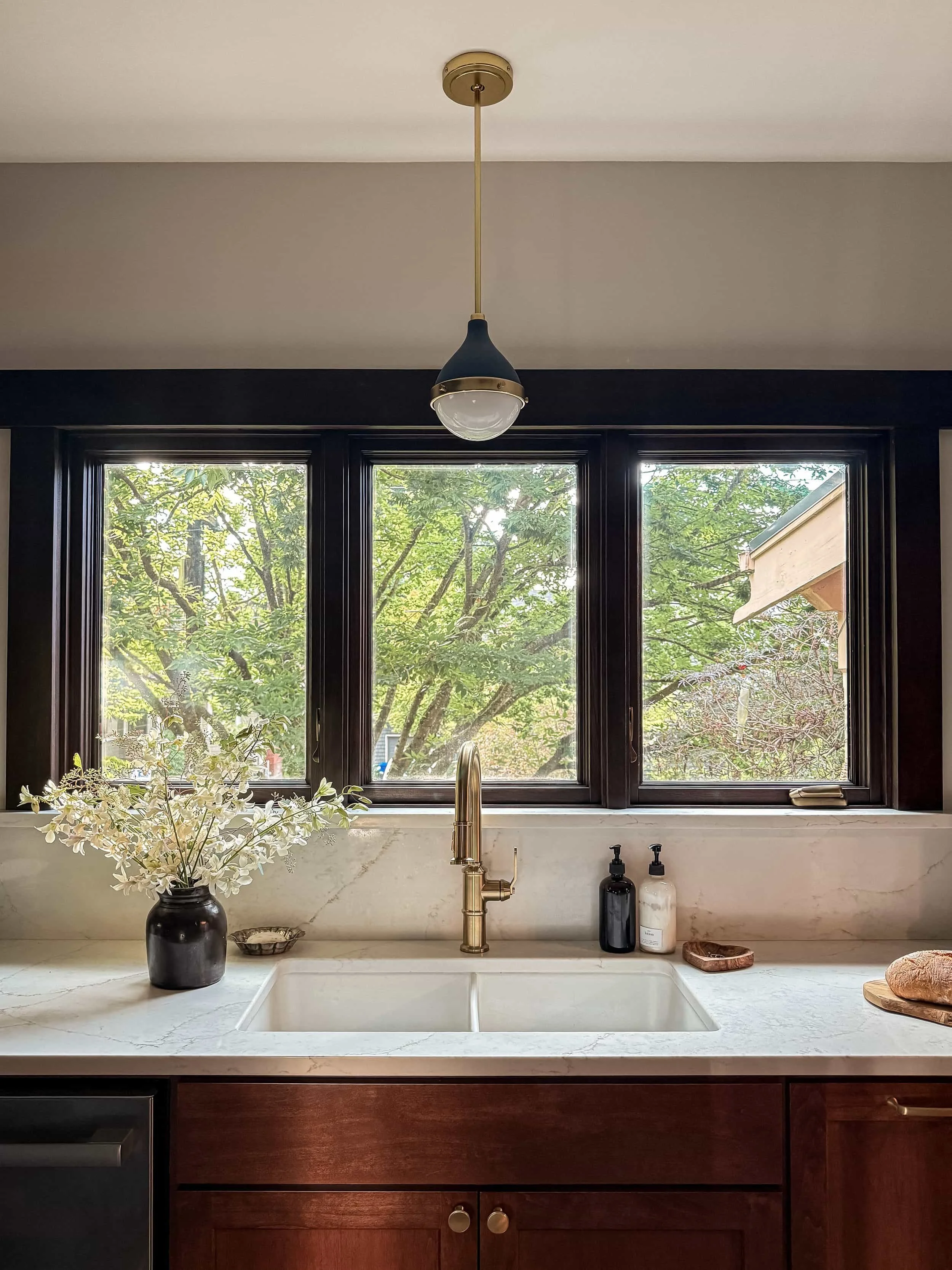 Kitchen sink area with a window view of trees outdoors, a black vase with white flowers, soap dispensers, and a wooden cutting board.