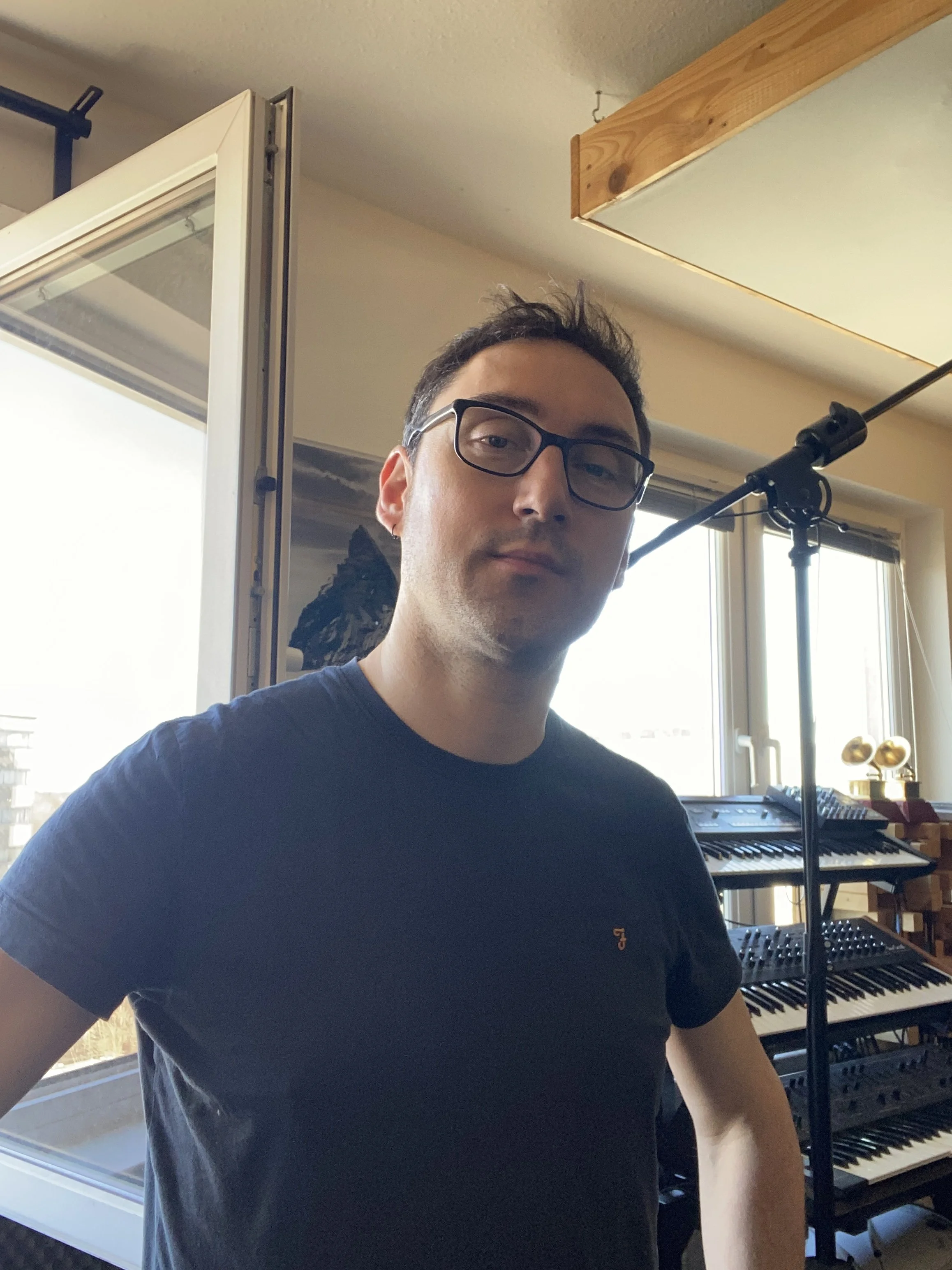 A man with glasses in a music studio, surrounded by musical equipment, standing near a window with sunlight coming in.
