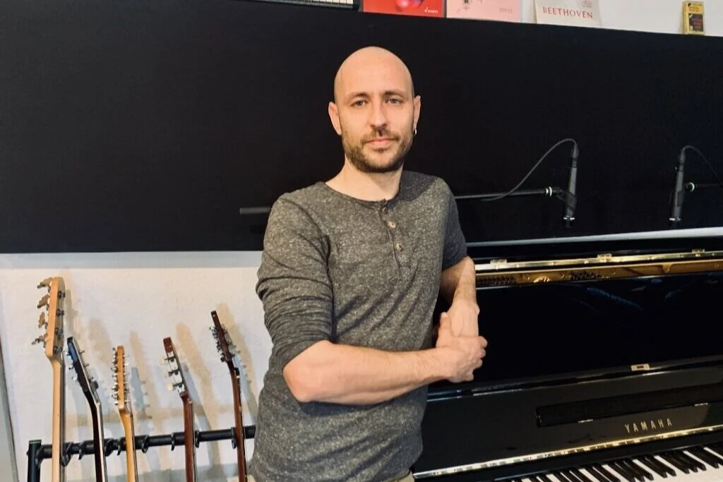 A man with a bald head and a beard standing next to a black Yamaha piano, with guitars on a stand behind him, in a music studio or room.