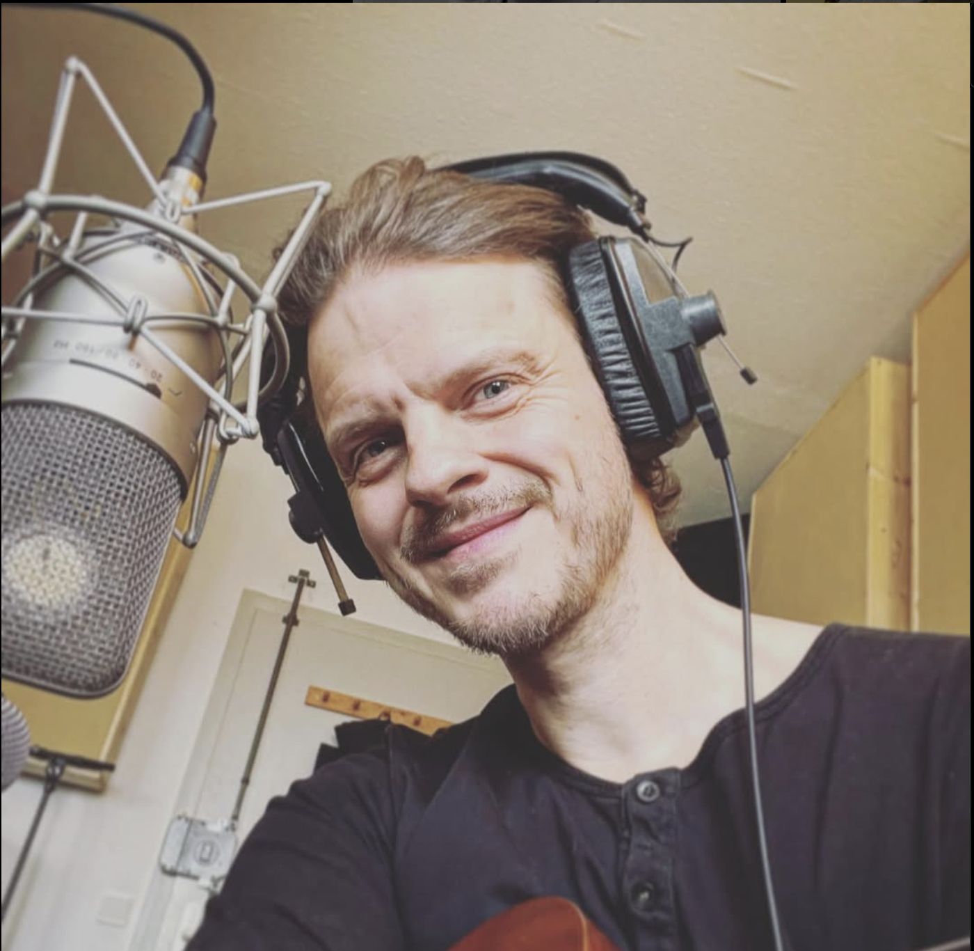 A man with short brown hair and a beard, wearing large headphones and a black shirt, is smiling at the camera in a recording studio with a professional microphone next to him.