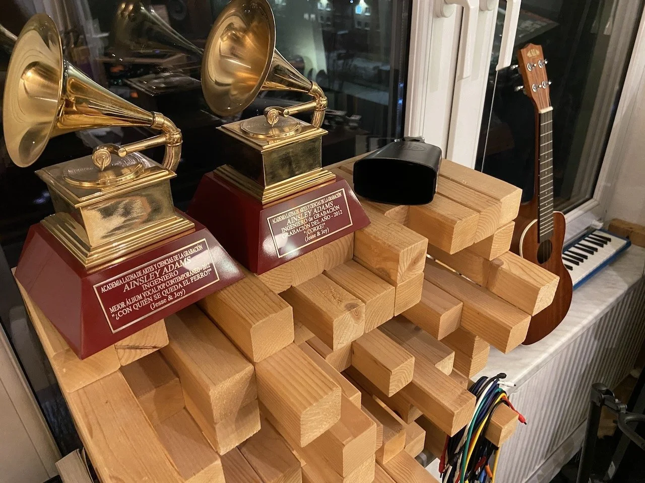 Two vintage gramophones with gold horns on wooden plaques with inscriptions, wooden blocks arranged in layered steps, a black speakers, a small guitar, a keyboard, and some colorful wires on a wooden table near a window.