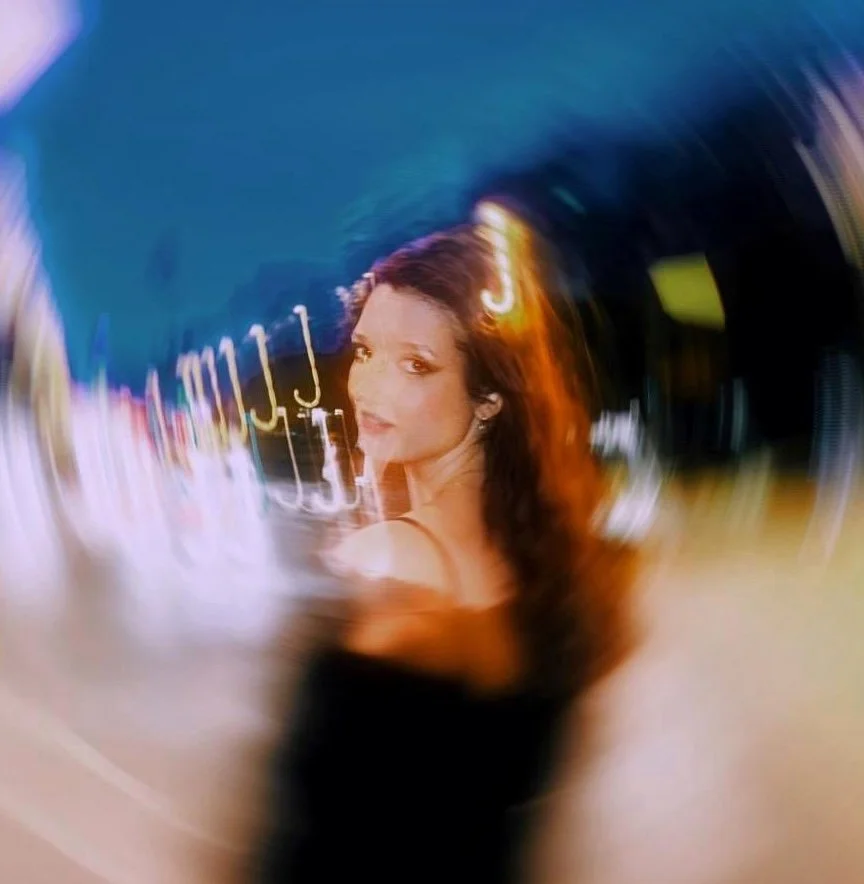 A woman with long, wavy brown hair looking at the camera, with blurred city lights and signs in the background seen through a swirl effect.
