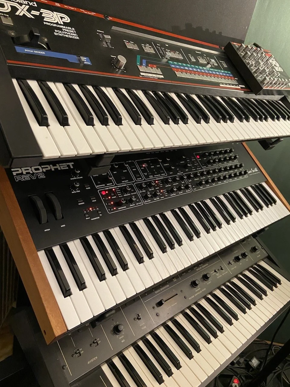 A three-tiered keyboard setup with synthesizers, including a Roland TR-808 drum machine on top, a Prophet Rev2 in the middle, and a Roland Juno-106 at the bottom.