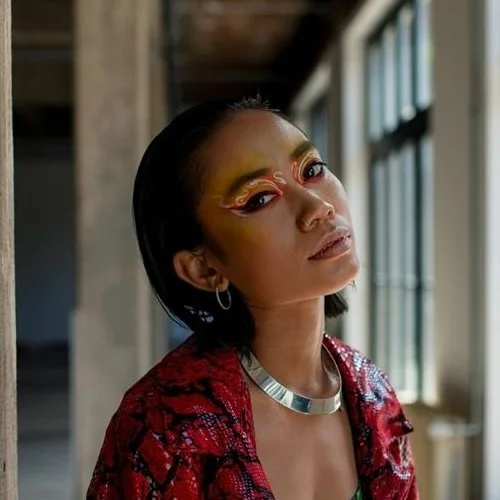 A woman with dramatic makeup, wearing a red patterned jacket and silver jewelry, looking at the camera inside a building with large windows.