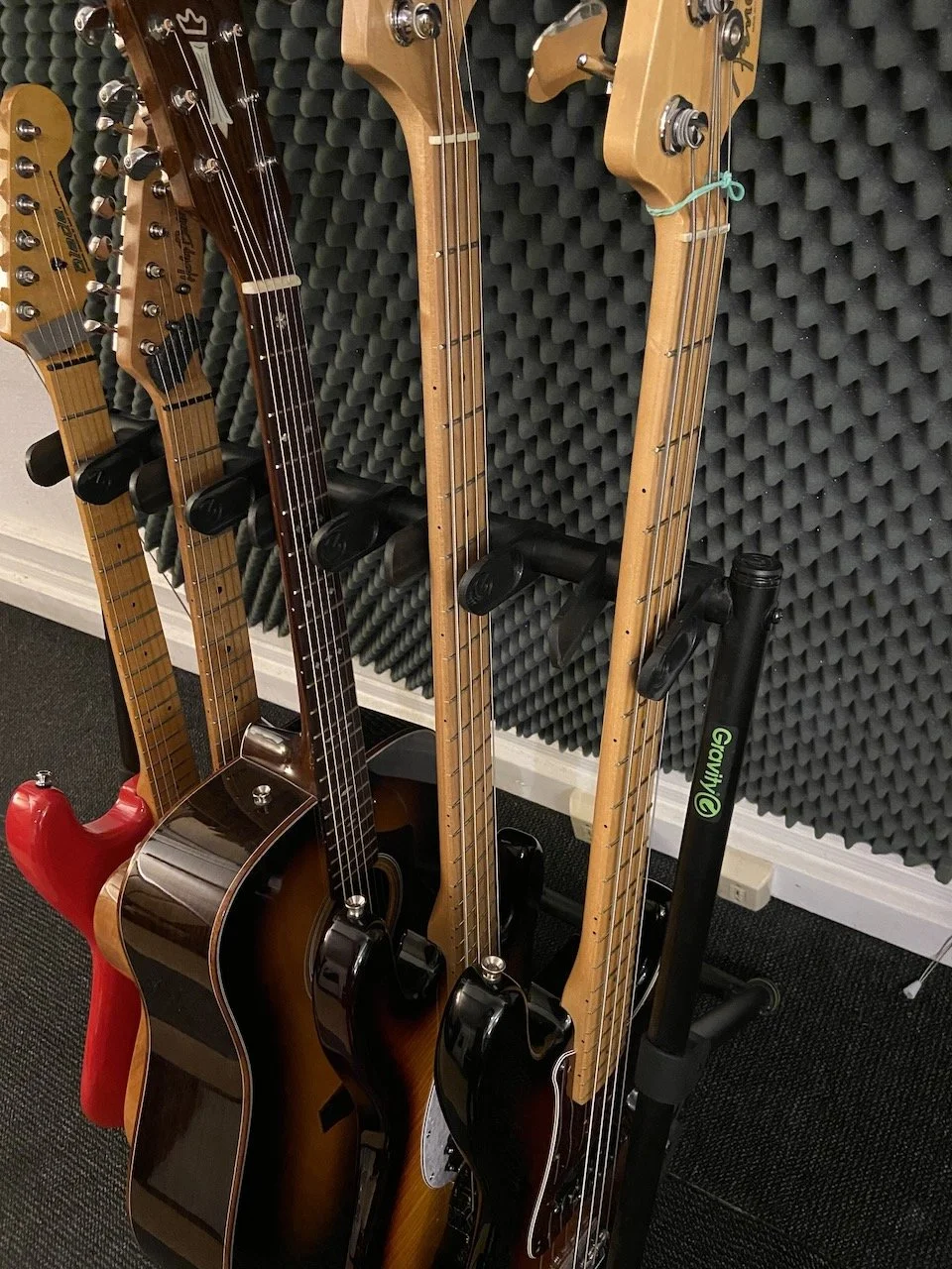 Collection of six guitars, including an acoustic, electric, and bass, resting on a guitar stand against soundproof foam panels.
