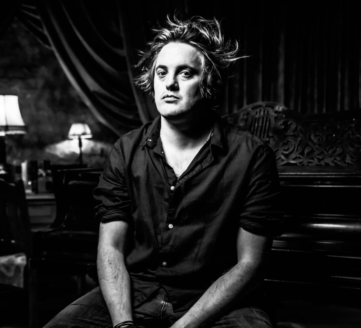 A young man with messy, shoulder-length hair, sitting in a dimly lit room, wearing a black shirt, looking directly at the camera. The background includes a piano, a lamp, and dark curtains.