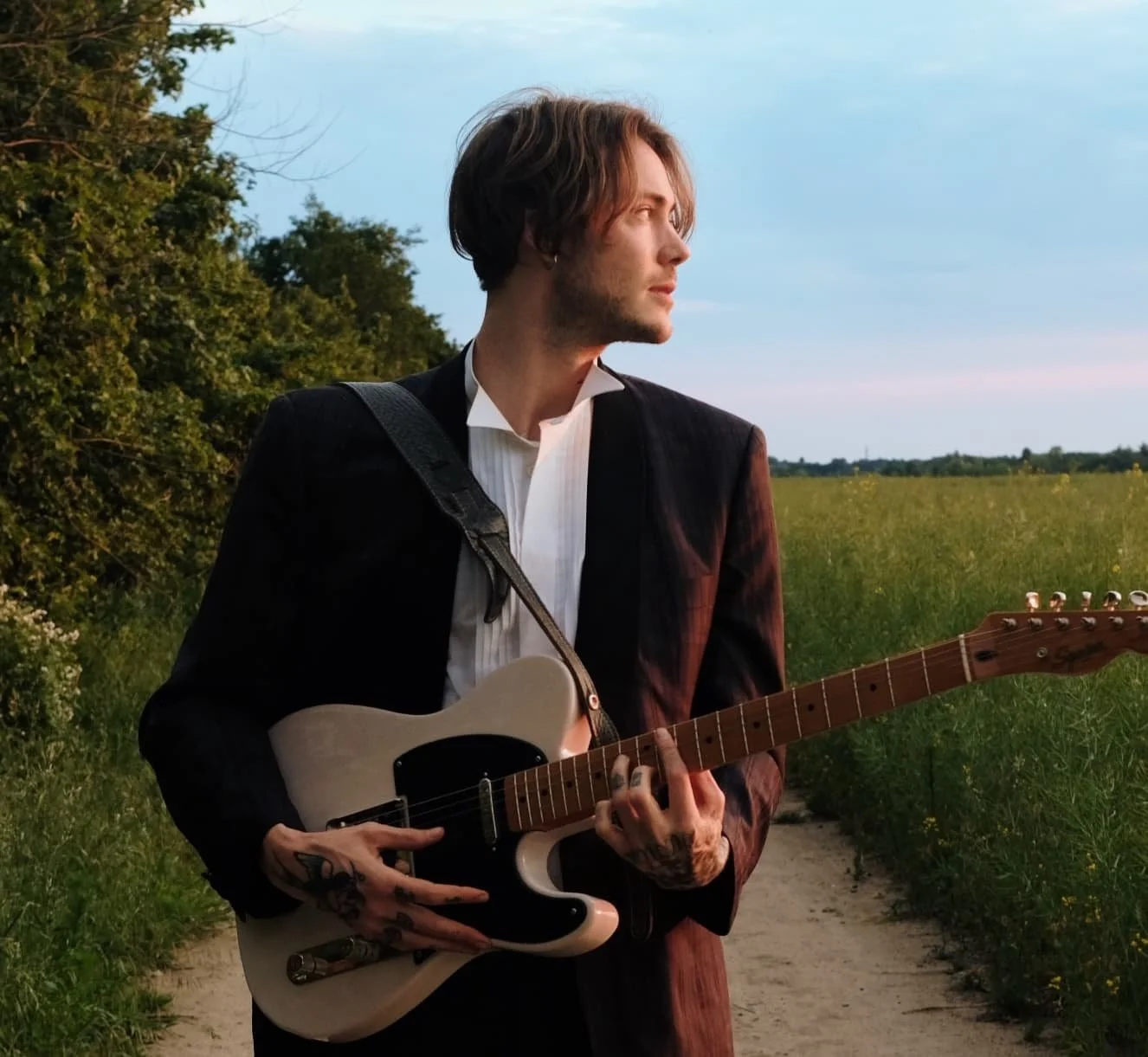 A young man with tattoos on his hands and light brown hair playing an electric guitar outdoors in a field during sunset.