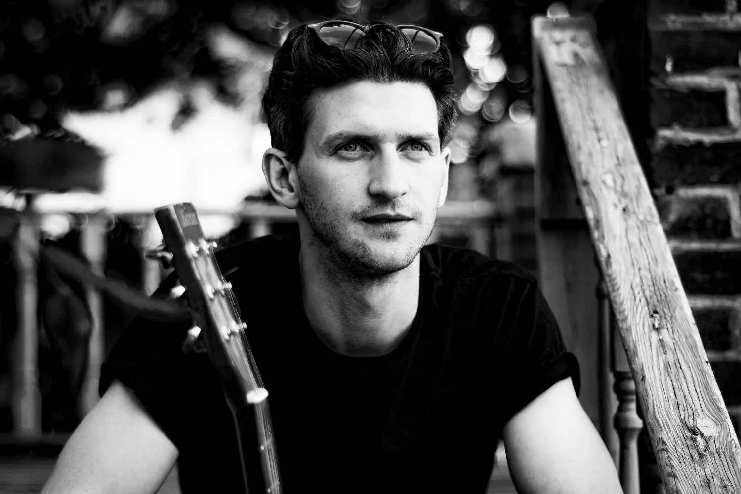 Black and white photo of a young man with dark hair, sitting outdoors, holding a guitar. He is looking off into the distance with a thoughtful expression, and there are sunglasses on his head. The background shows a wooden staircase and blurred trees.