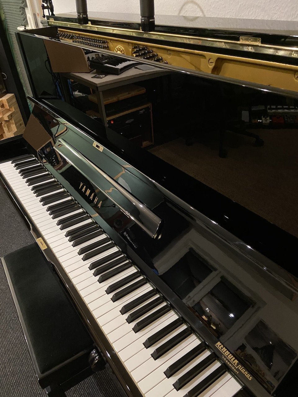 A black Yamaha upright piano in a music studio with a bench, with a reflection of the room visible on its polished surface. Another piano with an open lid and exposed strings is in the background.