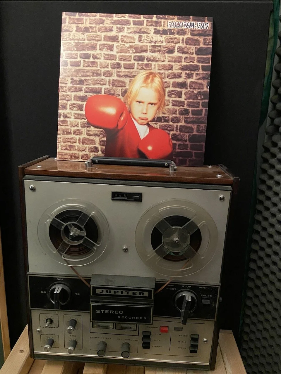 A vintage stereo recorder with spools and switches, placed in front of a vinyl album cover showing a child with boxing gloves aiming a punch.