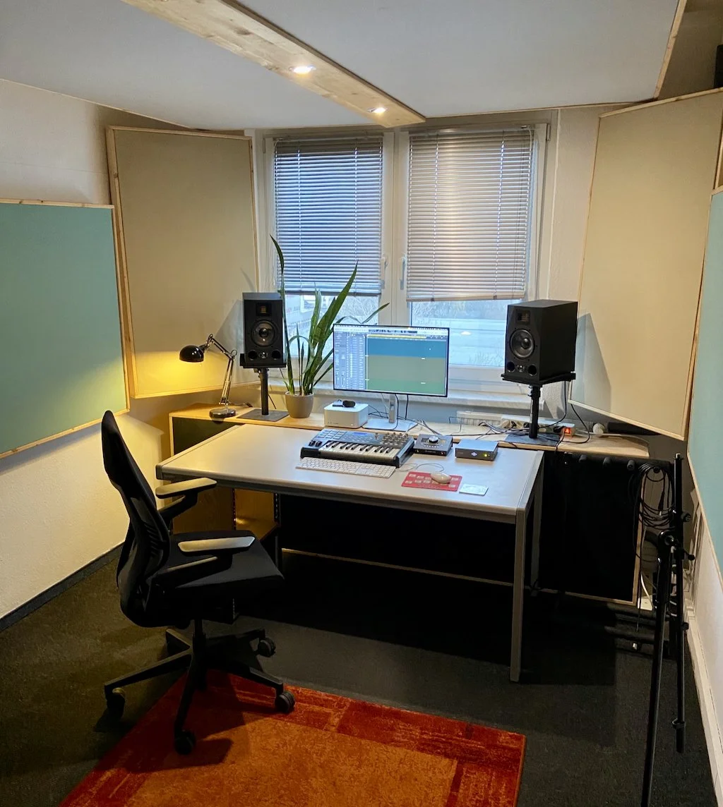 Recording studio or music production room with a desk, computer monitor, speakers, keyboard, mouse, microphone, and soundproofing on the walls and ceiling.