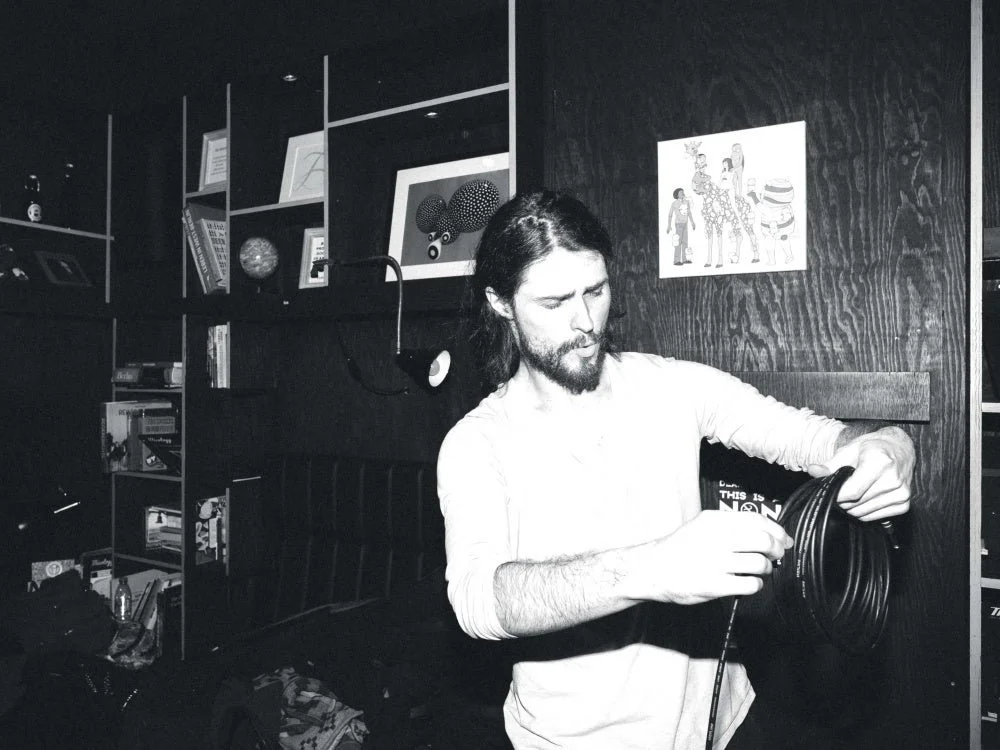 A man with long hair and a beard is holding and inspecting a coil of black cable in a room with dark wood paneling and bookshelf. There are artwork and books on the wall and shelves.
