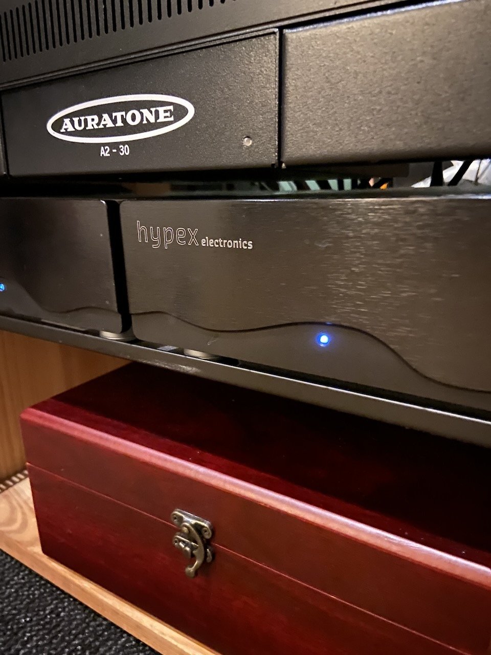 Close-up of electronic audio equipment with a black Auraton device and a black Hypex electronics device, placed on a wooden shelf below a wooden cabinet.
