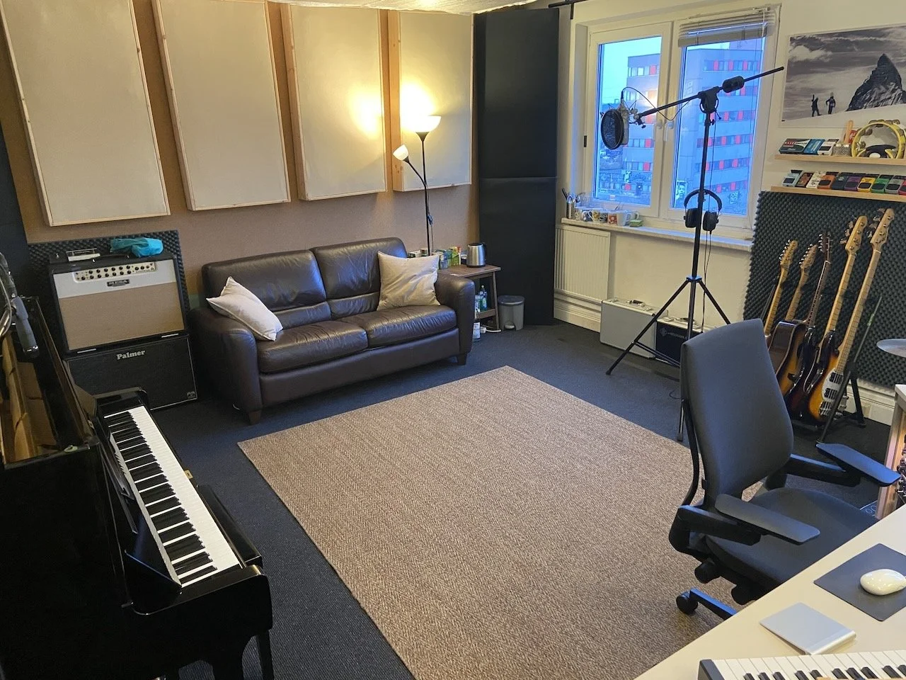 Recording studio with a black grand piano, a black leather sofa with two pillows, a tall floor lamp, music equipment including guitars on a stand, a microphone on a stand, a soundproof wall with acoustic panels, a window showing city buildings, and v