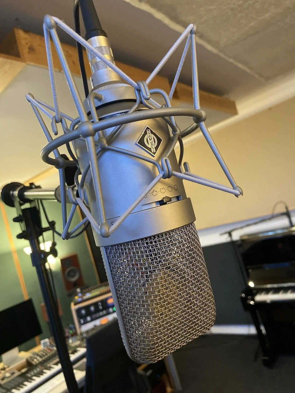 A studio microphone with a metallic grille and a shock mount, hanging in a recording studio, with musical instruments and sound equipment in the background.