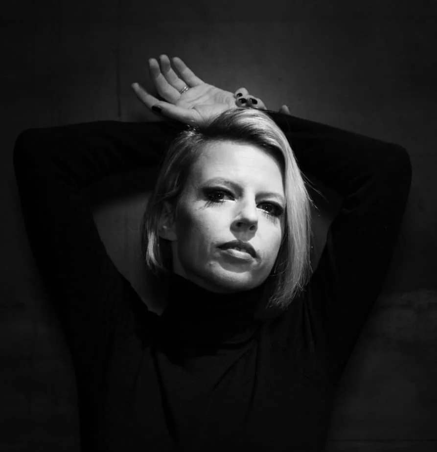 Black and white photo of a woman lying down with arms behind her head, looking at the camera.