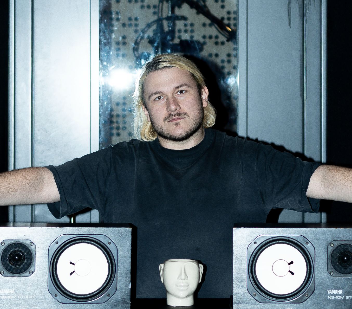 A man with long blonde hair, beard, and black t-shirt sitting in a music studio with two Yamaha studio monitor speakers on either side and a mannequin head between them.