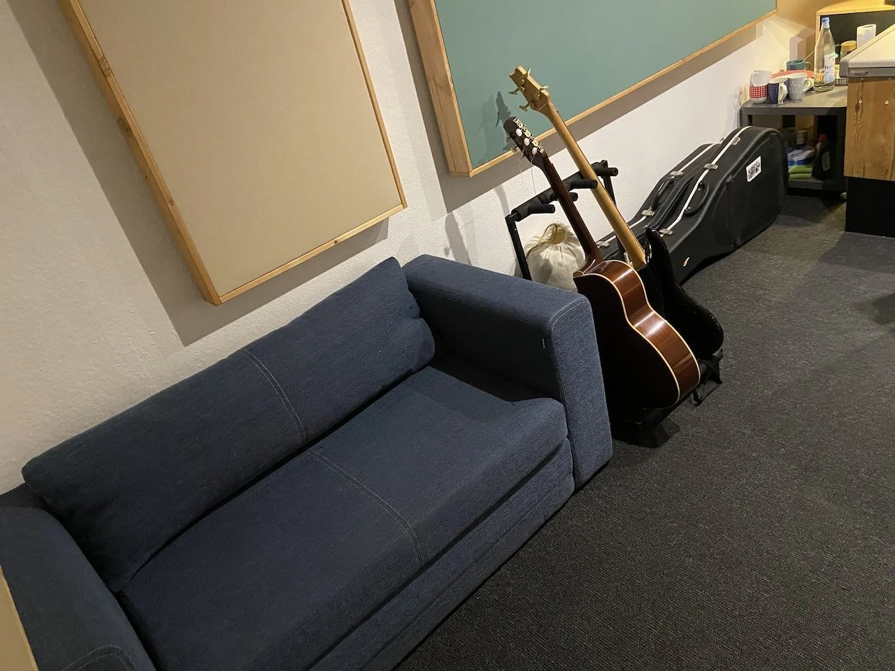A blue sofa with two guitars on a stand and a guitar case next to them in a room with beige and green soundproof panels on the wall.