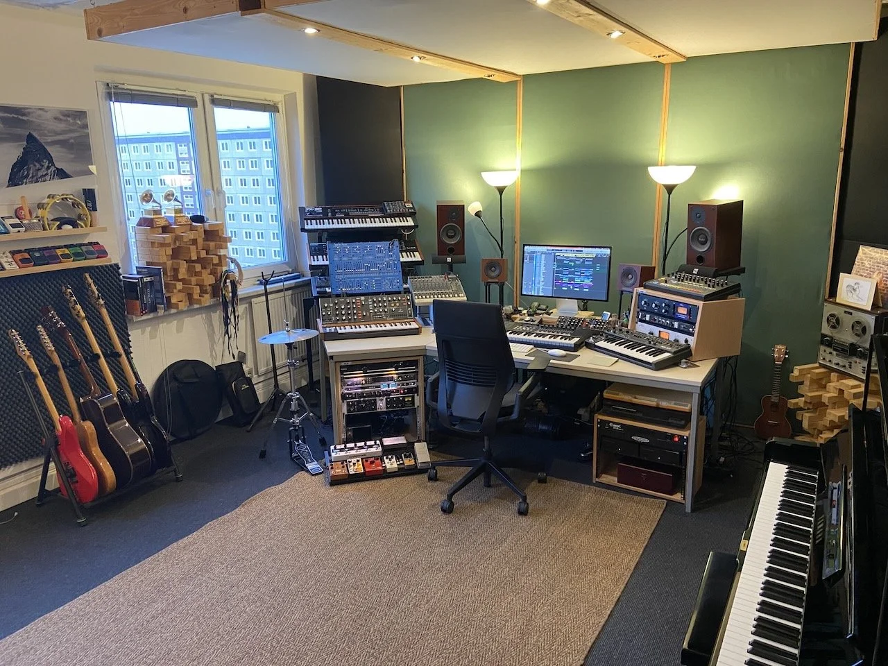 A music studio with a green wall, various guitars on a stand, a drum set, and multiple electronic music equipment including a keyboard, monitors, and mixing consoles. There are two lamps and acoustic treatment on the wall.