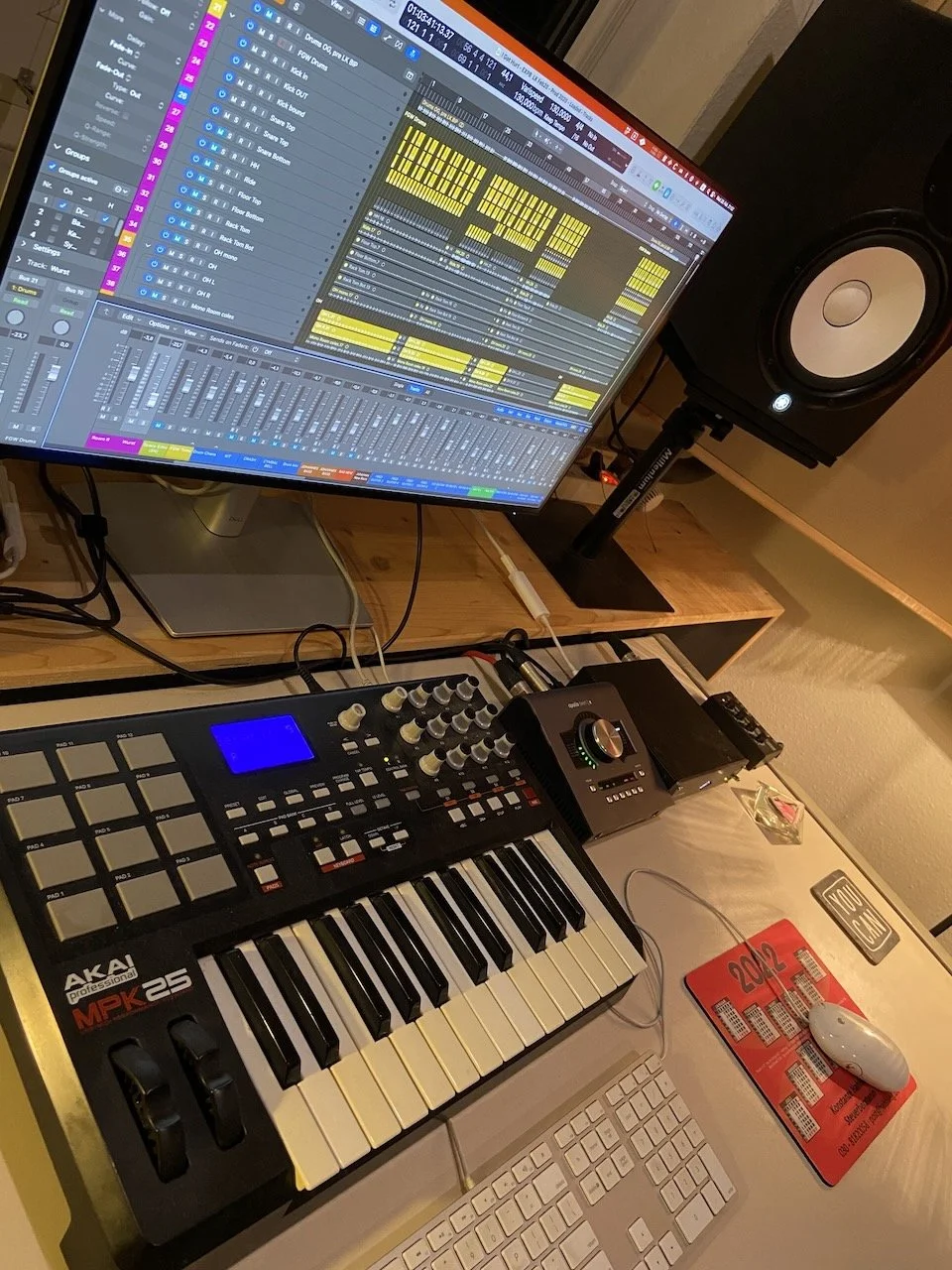 A music production workspace featuring a large computer monitor displaying digital audio workstation software, a MIDI keyboard, an audio interface, studio monitor speaker, a keyboard and mouse, and various audio equipment on a wooden desk.