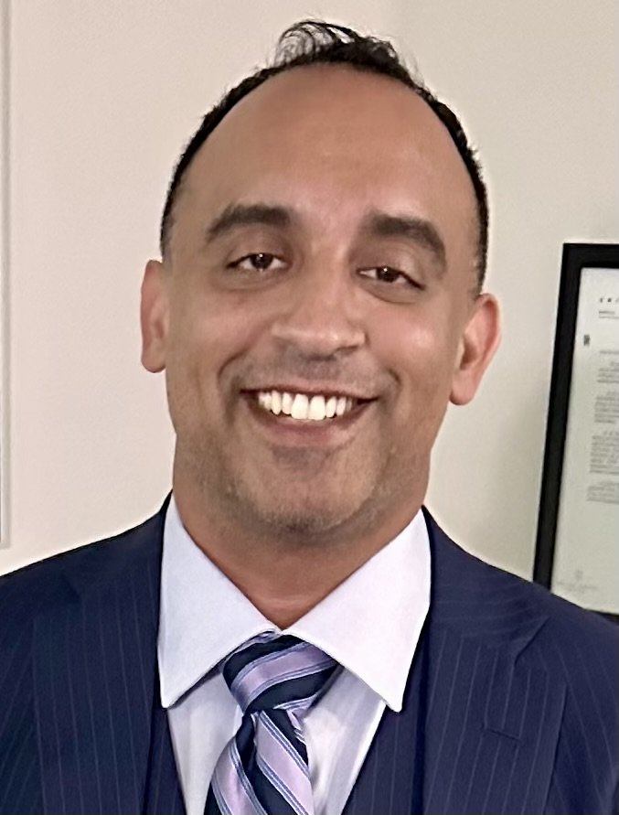 A man in a dark pinstripe suit, white shirt, and striped tie smiling at the camera.