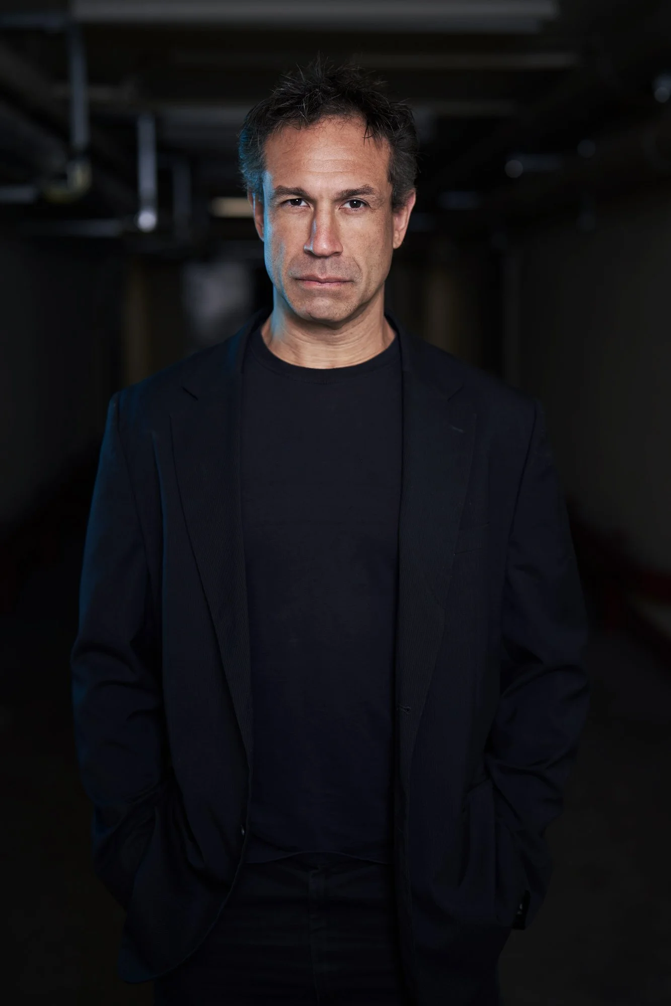 Porträtaufnahme von Evil Jared Hasselhoff (Bloodhound Gang) in schwarzem Blazer und Shirt, stehend in einem dunklen Kellergang, die Hände in den Taschen, kühles Seitenlicht, ernster Blick in die Kamera, fotografiert von Koszki Photography Berlin.