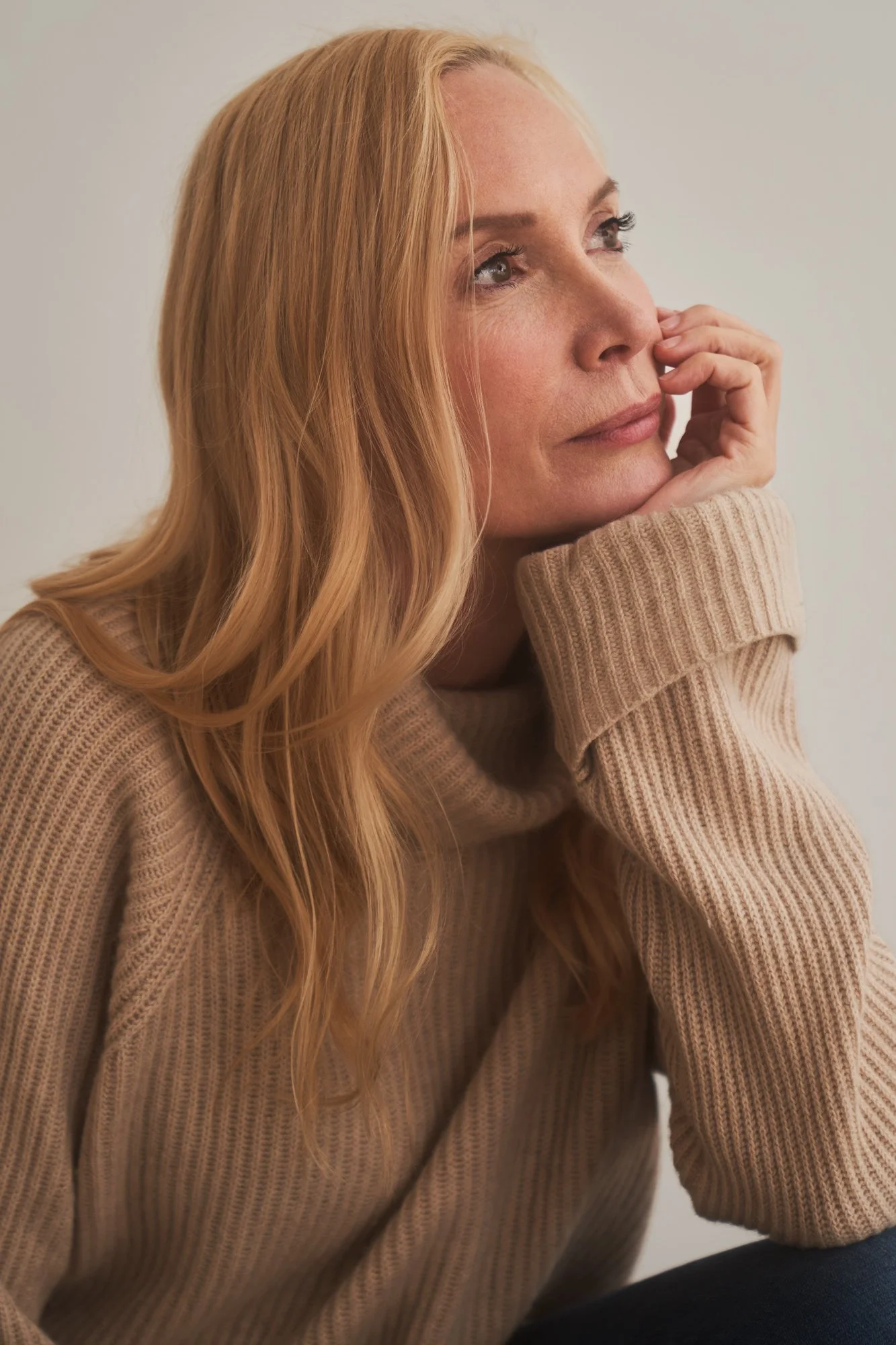 Seitenporträt der Regisseurin und Schauspielerin Feo Aladag mit langen blonden Haaren im beigen Strickpullover, das Kinn auf die Hand gestützt, mit nachdenklichem Blick zur Seite, fotografiert von Koszki Photography Berlin.