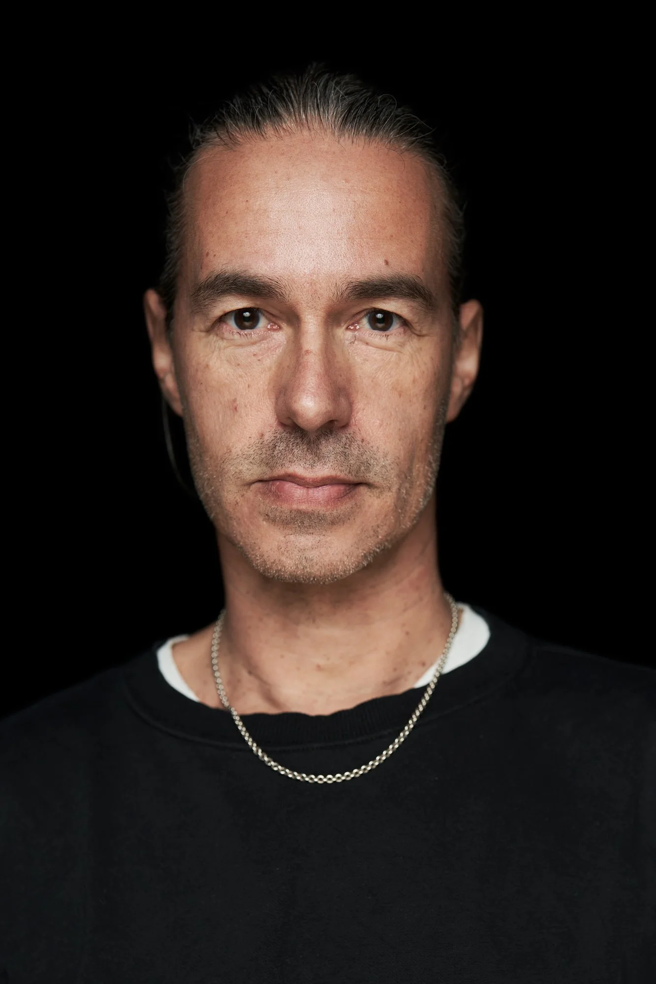Porträt des Gitarristen Christian Neander von der Rockband Selig, mit zurückgebundenen Haaren, silberner Kette und schwarzem Pullover vor schwarzem Hintergrund, fotografiert von Koszki Photography Berlin.