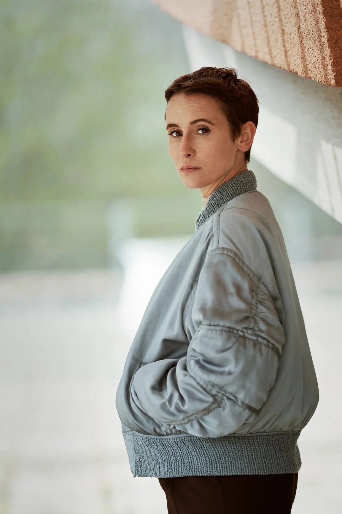 Porträtaufnahme der Schauspielerin Peri Baumeister in hellblauer Bomberjacke im Profil, Blick über die Schulter in die Kamera, vor Sichtbeton, fotografiert von Koszki Photography Berlin.