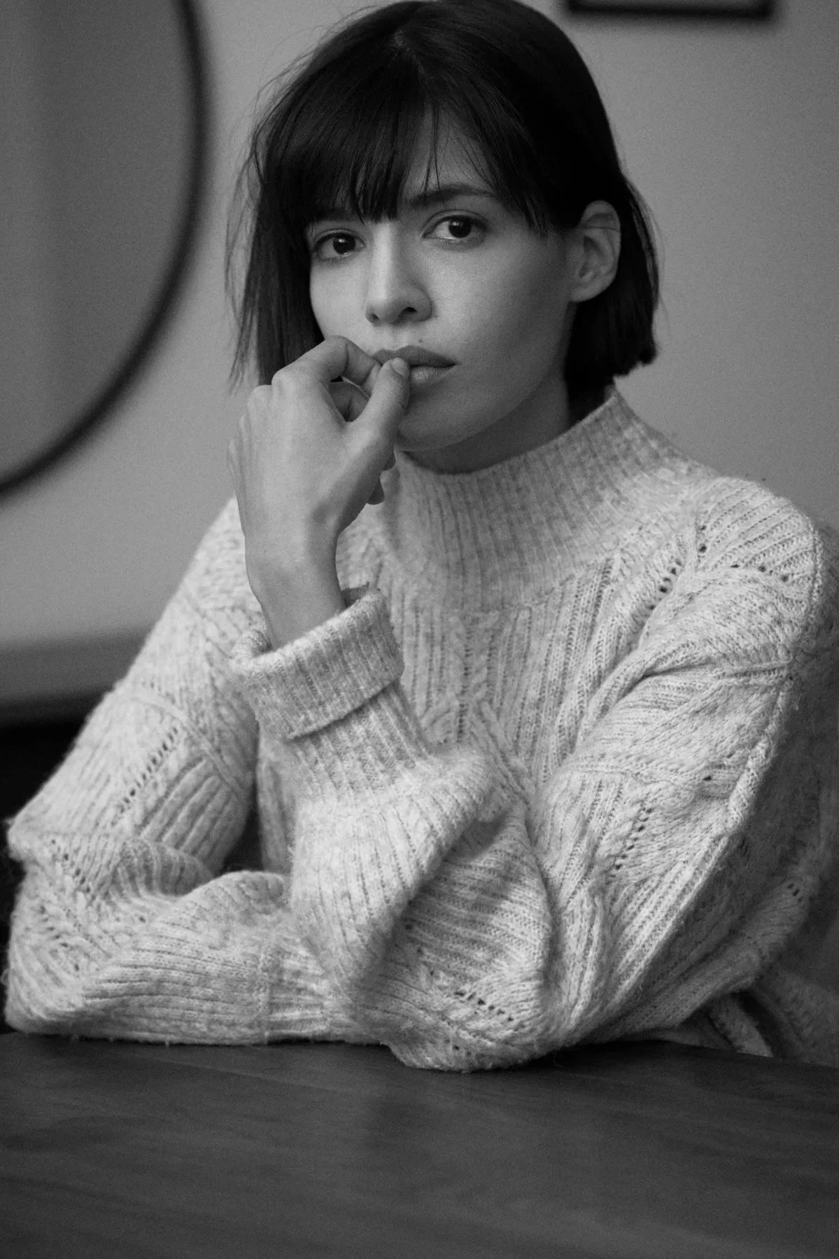 Schwarz-Weiß-Porträt der Schauspielerin Jasmin Lord im Strickpullover mit Stehkragen, am Tisch sitzend, die Hand an den Lippen, mit nachdenklichem Blick in die Kamera, fotografiert von Koszki Photography Berlin.