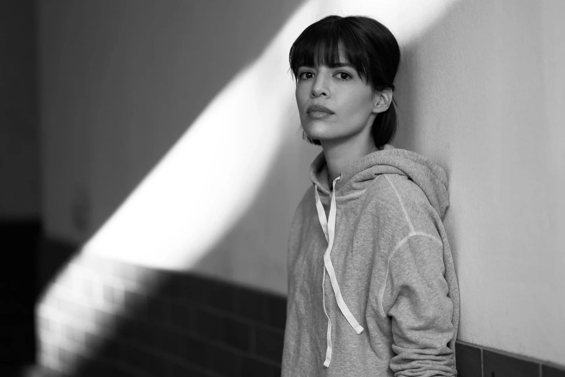 Schwarz-Weiß-Porträt der Schauspielerin Jasmin Lord im Kapuzenpullover, seitlich an eine Wand gelehnt, mit Blick über die Schulter in die Kamera im diagonalen Lichteinfall eines Treppenhauses, fotografiert von Koszki Photography Berlin.
