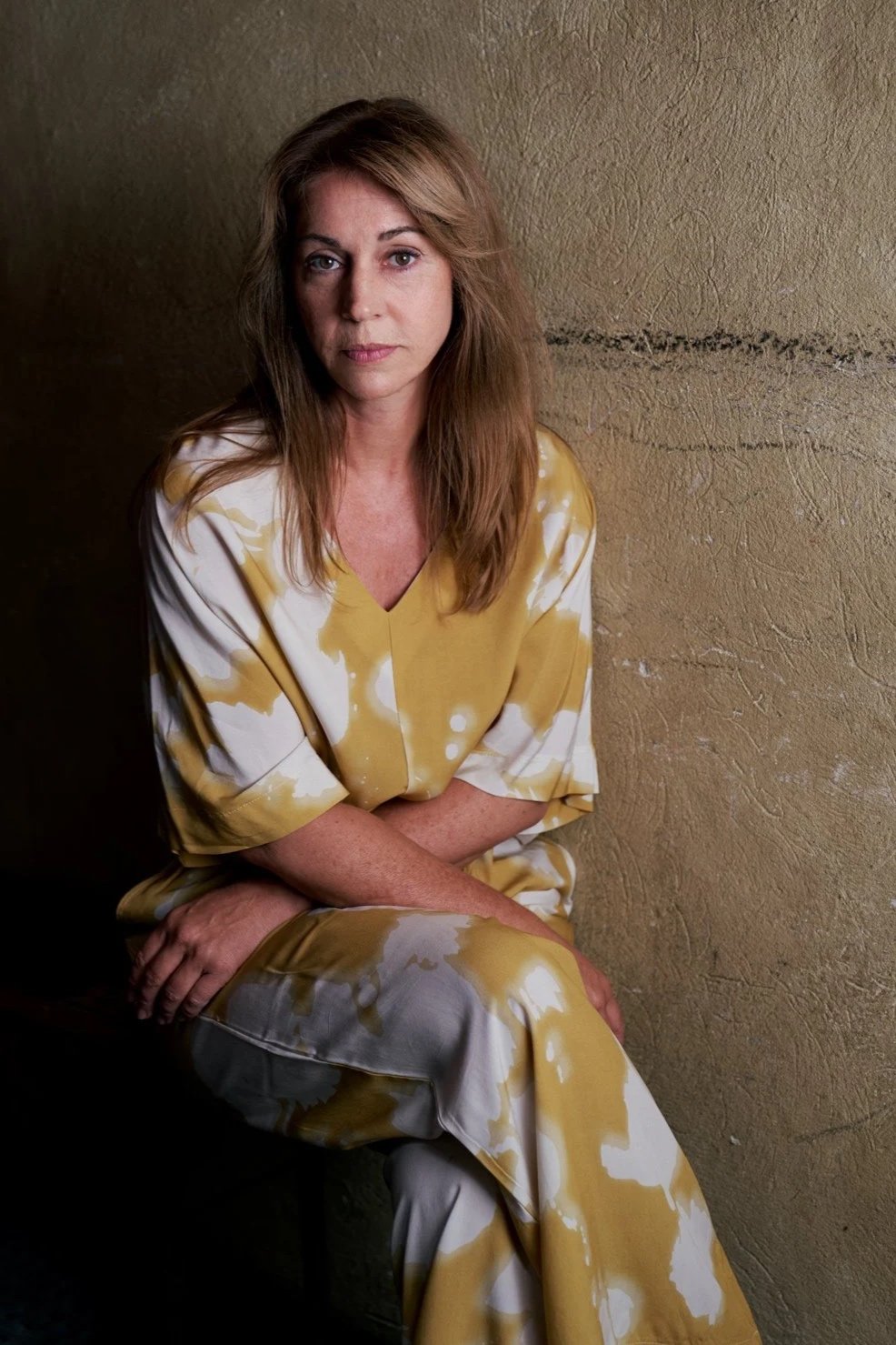 Porträtaufnahme der Schauspielerin Ulrike C. Tscharre im senfgelb-weiß gemusterten Kleid, sitzend vor einer strukturierten ockerfarbenen Wand, ruhiger Blick in die Kamera, fotografiert von Koszki Photography Berlin.