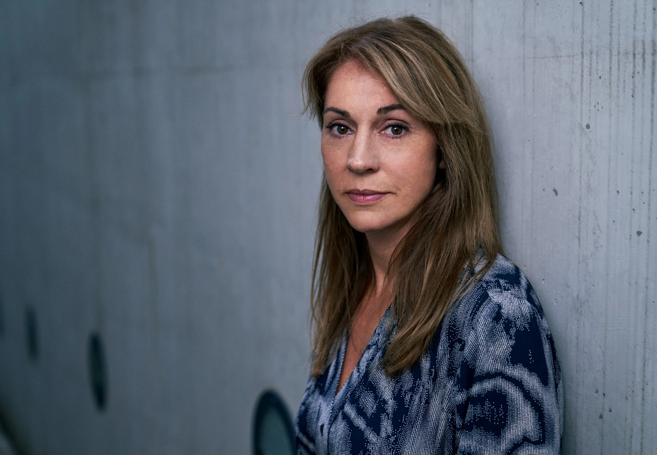 Porträtaufnahme der Schauspielerin Ulrike C. Tscharre im blau-grau gemusterten Kleid, seitlich an eine Betonwand gelehnt, Blick über die Schulter in die Kamera, fotografiert von Koszki Photography Berlin.
