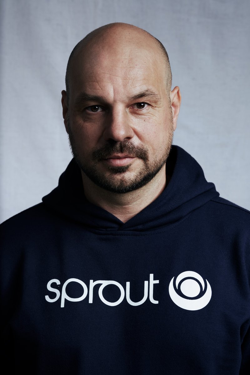 Porträt des DJs D-Nox im dunkelblauen Sprout-Hoodie vor hellgrauem Hintergrund mit ernstem Blick, fotografiert von Koszki Photography Berlin.