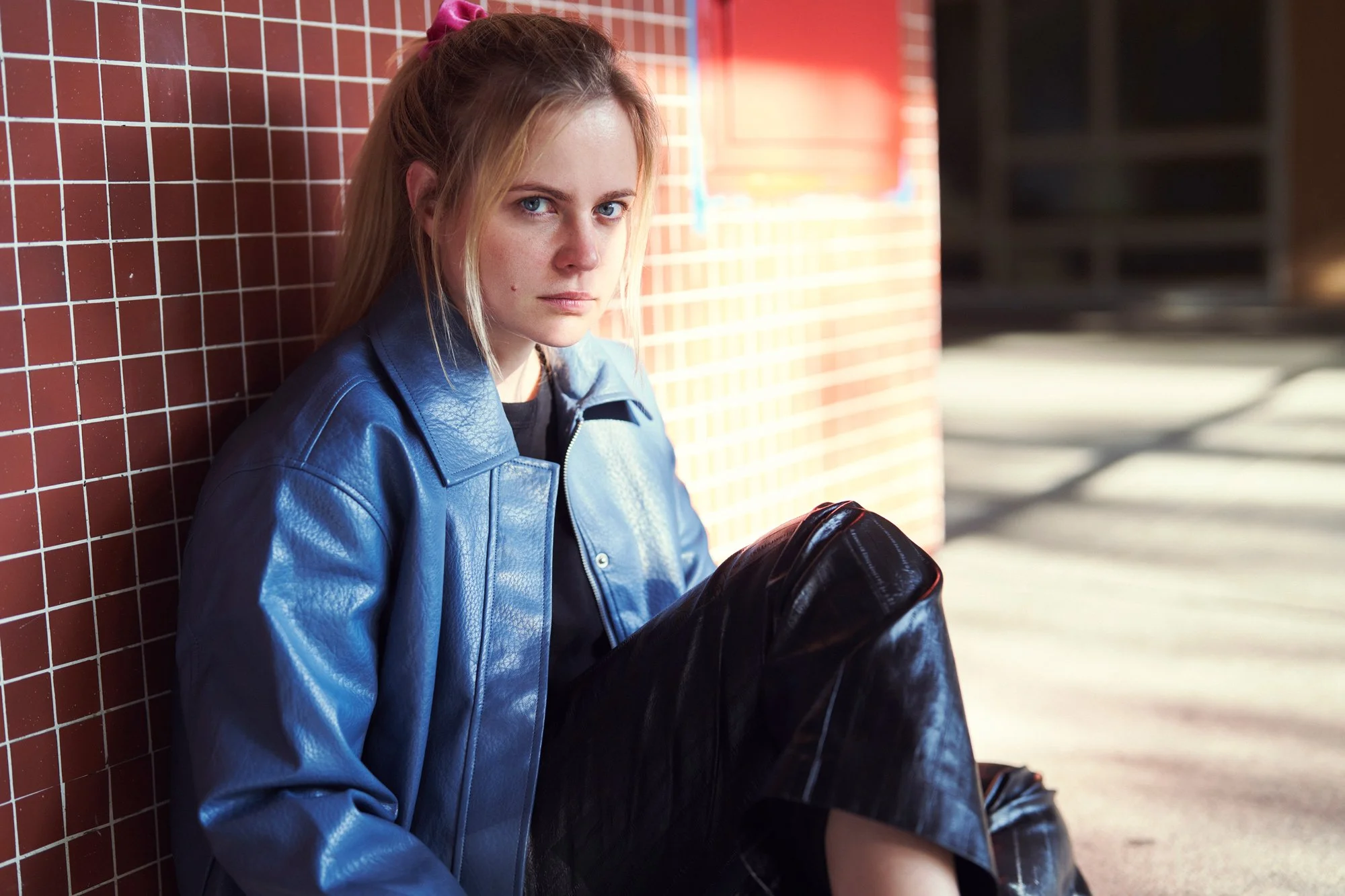 Porträt der Schauspielerin und Sängerin Kara Edenberg im blauen Lederjackett und schwarzer Lederhose, mit rosa Haargummi am Boden sitzend und an eine rote Kachelwand gelehnt, mit ernstem Blick in die Kamera im harten Sonnenlicht, fotografiert von Kos