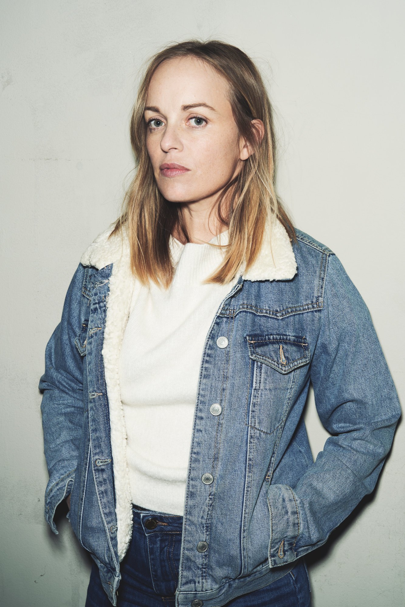 Porträtaufnahme der Schauspielerin Friederike Kempter in Jeansjacke mit Teddyfutter-Kragen und weißem Pullover vor heller Wand, fotografiert von Koszki Photography Berlin.