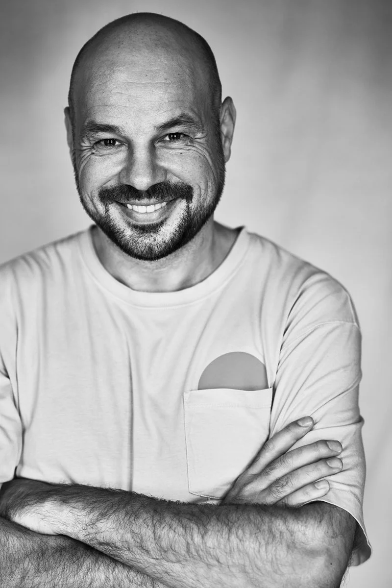 Schwarz-Weiß-Porträt des DJs D-Nox, lächelnd mit verschränkten Armen vor hellem Hintergrund, fotografiert von Koszki Photography Berlin.