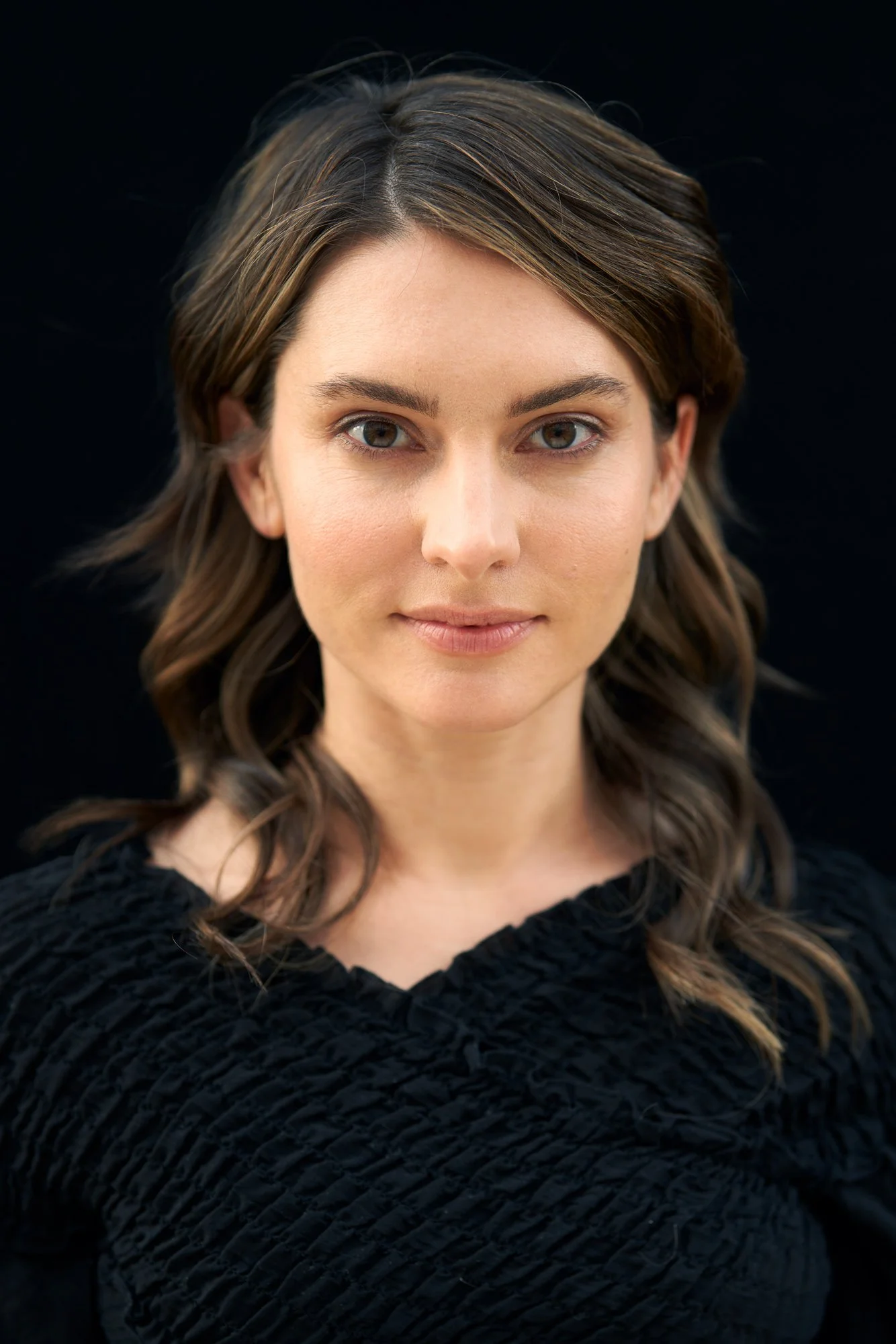 Headshot der Schauspielerin Romina Küper mit welligem dunklem Haar in schwarzem Oberteil mit Raffung vor schwarzem Hintergrund, frontaler Blick in die Kamera, fotografiert von Koszki Photography Berlin.