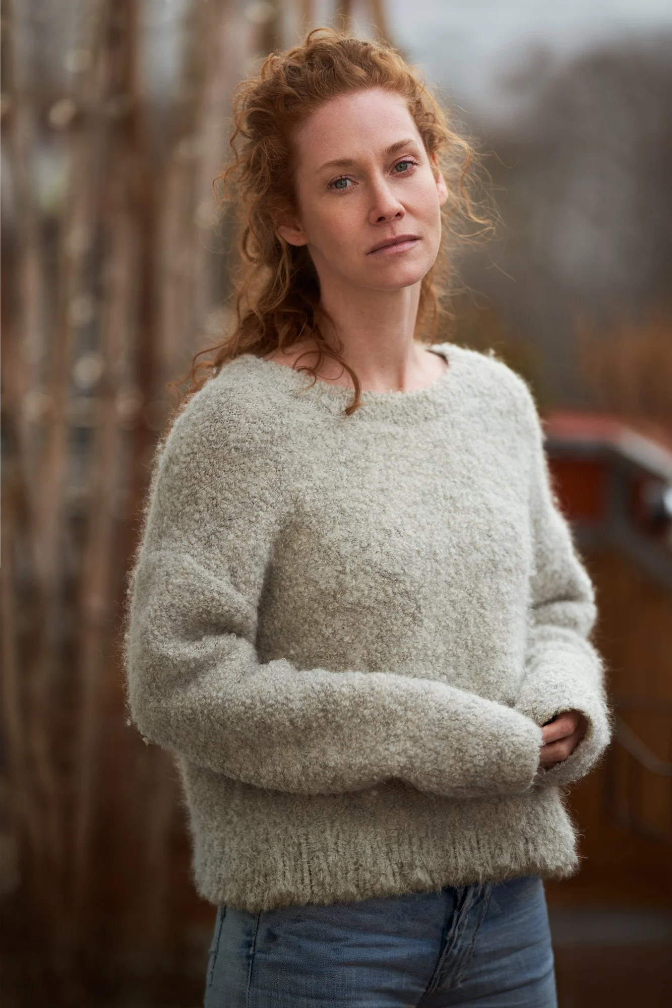 Porträtaufnahme der Schauspielerin Sina-Valeska Jung mit lockigem rotem Haar in grauem Strickpullover und Jeans, draußen im weichen Tageslicht, nachdenklicher Blick, fotografiert von Koszki Photography Berlin.